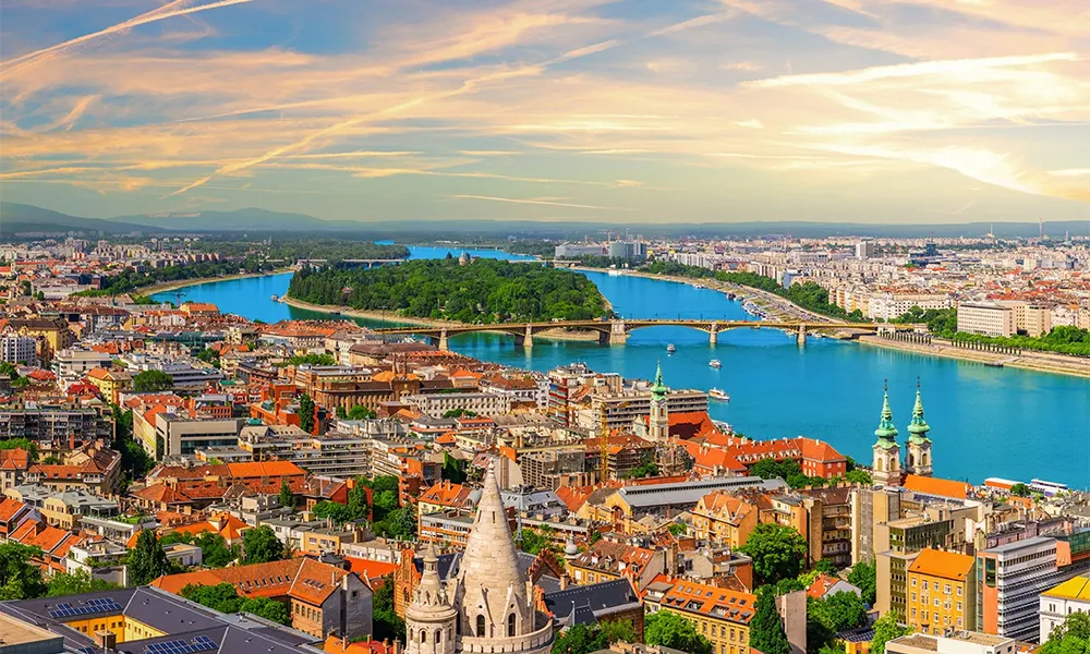 An aerial view of Budapest, Hungary, with the Danube river winding past the city skyline at sunset.